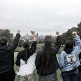 Five people standing outdoors with their backs to the camera, raising fists in the air, with a city skyline visible in the distance.